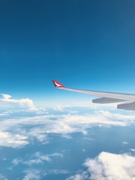 An airplane wing extends into the sky, with a clear view of fluffy white clouds below and a bright blue sky above. The wing tip shows a red marking or logo.