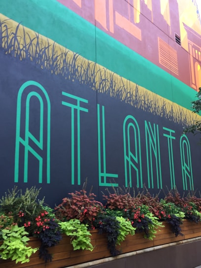A mural featuring the word 'ATLANTA' in large, stylized green letters. Above the word, there's an artistic design resembling city buildings and vegetation. A planter box is filled with diverse foliage and flowers in shades of green, red, and purple, adding natural contrast to the mural.