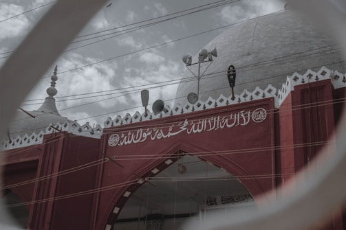 A red building with ornate white Arabic script on its façade and a large dome on top. The structure features several antennas and loudspeakers, with a cloudy sky in the background. There are birds flying near the dome, and the scene is viewed through a circular frame.