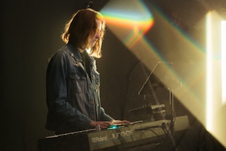 A person is playing a Roland JUNO-DS keyboard in a dimly lit environment. They wear a denim jacket and are illuminated by a beam of light that creates a prism effect, casting colorful reflections.