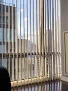 Vertical blinds partially cover a large window displaying a view of modern high-rise buildings against a blue sky with scattered clouds. Reflections and shadows are cast on the wooden floor, adding a sense of depth.