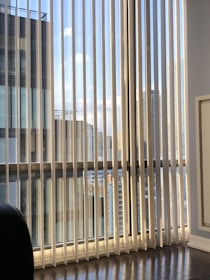 Vertical blinds partially cover a large window displaying a view of modern high-rise buildings against a blue sky with scattered clouds. Reflections and shadows are cast on the wooden floor, adding a sense of depth.