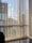 Vertical blinds partially cover a large window displaying a view of modern high-rise buildings against a blue sky with scattered clouds. Reflections and shadows are cast on the wooden floor, adding a sense of depth.