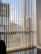 Vertical blinds partially cover a large window displaying a view of modern high-rise buildings against a blue sky with scattered clouds. Reflections and shadows are cast on the wooden floor, adding a sense of depth.