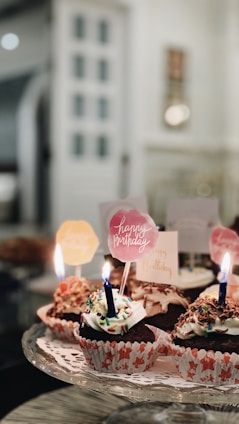 A selection of cupcakes with colorful frosting and sprinkles, each topped with a lit birthday candle. The cupcakes are arranged on a decorative serving tray. In the background, there are blurred images of a room interior, suggesting a casual, cozy setting.