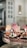 A selection of cupcakes with colorful frosting and sprinkles, each topped with a lit birthday candle. The cupcakes are arranged on a decorative serving tray. In the background, there are blurred images of a room interior, suggesting a casual, cozy setting.