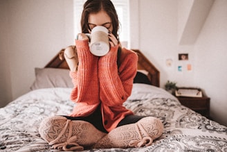 A person is sitting cross-legged on a bed, wearing a cozy knit sweater and patterned leggings, holding a mug close to their face. The bedroom features a wooden headboard and patterned bedspread. There is a typewriter and some photos on the wall in the background.
