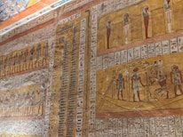 Ancient Egyptian wall art featuring hieroglyphics and depictions of figures in traditional poses and garments. The art includes multiple registers of figures, some with headdresses, surrounded by text. The background primarily consists of ochre and earth tones, with vibrant blue and red accents.