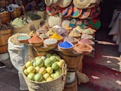 A vibrant market stall displays an array of spices in a variety of colors, arranged in small bowls on a wooden surface. Behind the spices, there's a decorative backdrop of woven baskets with colorful patterns. In the foreground, a basket full of lemons adds to the lively atmosphere.