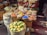 A vibrant market stall displays an array of spices in a variety of colors, arranged in small bowls on a wooden surface. Behind the spices, there's a decorative backdrop of woven baskets with colorful patterns. In the foreground, a basket full of lemons adds to the lively atmosphere.