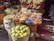 A vibrant market stall displays an array of spices in a variety of colors, arranged in small bowls on a wooden surface. Behind the spices, there's a decorative backdrop of woven baskets with colorful patterns. In the foreground, a basket full of lemons adds to the lively atmosphere.