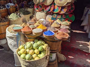 A vibrant market stall displays an array of spices in a variety of colors, arranged in small bowls on a wooden surface. Behind the spices, there's a decorative backdrop of woven baskets with colorful patterns. In the foreground, a basket full of lemons adds to the lively atmosphere.