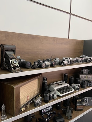 A collection of vintage cameras and equipment is displayed on wooden shelves. The top shelf features a lineup of old cameras including a large format camera and several SLR models. The middle shelf showcases a retro radio, a typewriter, and a variety of camera lenses. Additional shelves have more cameras and accessories, creating a nostalgic display of antique photography equipment.