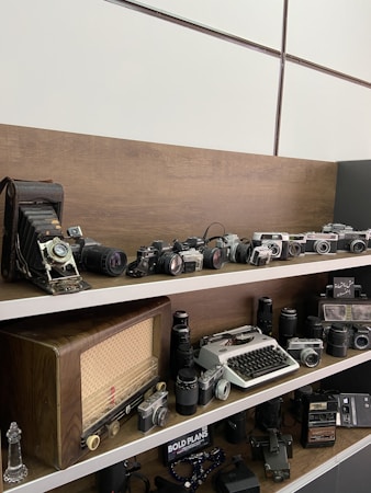 A collection of vintage cameras and equipment is displayed on wooden shelves. The top shelf features a lineup of old cameras including a large format camera and several SLR models. The middle shelf showcases a retro radio, a typewriter, and a variety of camera lenses. Additional shelves have more cameras and accessories, creating a nostalgic display of antique photography equipment.