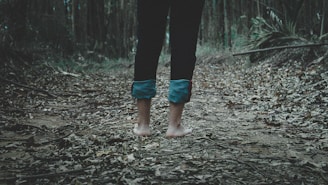 Barefoot person standing on a path covered with dry leaves in a dense forest. The legs are partially visible, clad in dark pants rolled up at the bottom. The surroundings are filled with trees and scattered foliage.