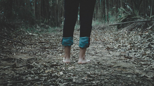 Barefoot person standing on a path covered with dry leaves in a dense forest. The legs are partially visible, clad in dark pants rolled up at the bottom. The surroundings are filled with trees and scattered foliage.