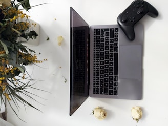 A silver laptop is open on a white surface, with a black game controller resting on its right side. To the left, a bouquet of lush greenery and yellow flowers sits, with a couple of white roses placed on the table nearby.