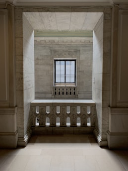 An elegant space with marble walls and a decorative balustrade. A window with a grid design is centered in the background, allowing soft light to enter the room, creating a calm and serene atmosphere.