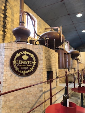 A rustic distillery interior with large copper stills and brick walls, featuring an ornate sign for 'Lepanto Gonzalez Byass Xerez.' The setup includes a long corridor with a red railing and bottles placed on round red tables.