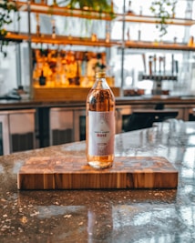 A bottle of rosé wine with a label reading 'Forty Ounce Rosé' sits on a wooden cutting board on a polished marble or granite countertop. In the background, there are shelves with various bottles and plants, providing a warm and inviting atmosphere.