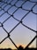 A chain-link fence is silhouetted against a sky transitioning from light blue to a deeper shade, with a small white moon visible through the gaps. The horizon shows a faint sunset with soft orange and yellow tones, with silhouettes of trees and light poles in the distance.