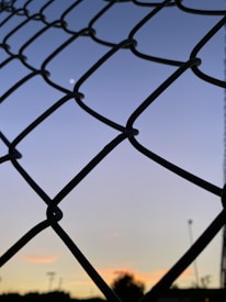 A chain-link fence is silhouetted against a sky transitioning from light blue to a deeper shade, with a small white moon visible through the gaps. The horizon shows a faint sunset with soft orange and yellow tones, with silhouettes of trees and light poles in the distance.