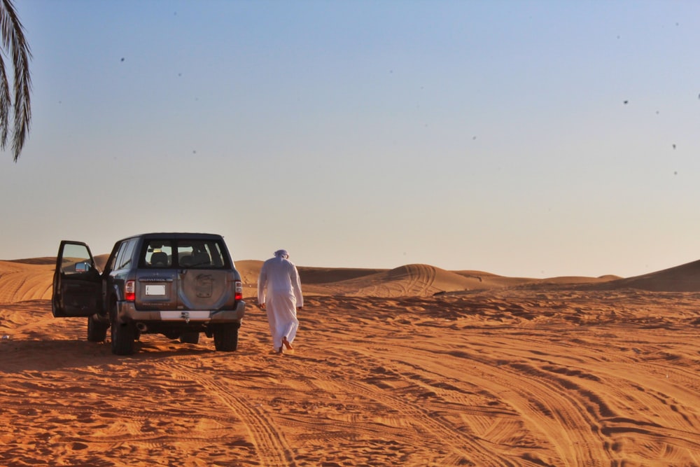 Mann im weißen Langarmhemd steht tagsüber neben schwarzem SUV auf braunem Sand