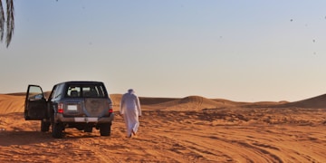 man in white long sleeve shirt standing beside black suv on brown sand during daytime