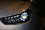 Close-up of a modern car headlight displaying a sleek and aerodynamic design. The headlight features LED technology with a clean and polished appearance. The car body is finished in a metallic color, reflecting light subtly.