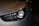 Close-up of a modern car headlight displaying a sleek and aerodynamic design. The headlight features LED technology with a clean and polished appearance. The car body is finished in a metallic color, reflecting light subtly.