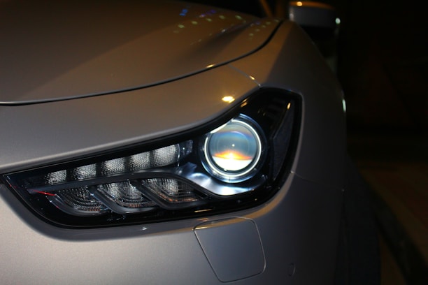 Close-up of a modern car headlight displaying a sleek and aerodynamic design. The headlight features LED technology with a clean and polished appearance. The car body is finished in a metallic color, reflecting light subtly.