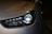 Close-up of a modern car headlight displaying a sleek and aerodynamic design. The headlight features LED technology with a clean and polished appearance. The car body is finished in a metallic color, reflecting light subtly.