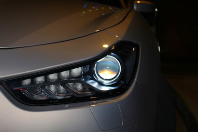 Close-up of a modern car headlight displaying a sleek and aerodynamic design. The headlight features LED technology with a clean and polished appearance. The car body is finished in a metallic color, reflecting light subtly.