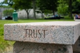 A stone bench with the word 'TRUST' engraved on its side is set in a lush, green park. The background features trees, blurred foliage, and a few indistinct people, creating a serene and calming atmosphere.