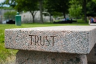A stone bench with the word 'TRUST' engraved on its side is set in a lush, green park. The background features trees, blurred foliage, and a few indistinct people, creating a serene and calming atmosphere.