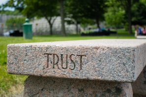 A stone bench with the word 'TRUST' engraved on its side is set in a lush, green park. The background features trees, blurred foliage, and a few indistinct people, creating a serene and calming atmosphere.