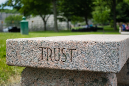 A stone bench with the word 'TRUST' engraved on its side is set in a lush, green park. The background features trees, blurred foliage, and a few indistinct people, creating a serene and calming atmosphere.