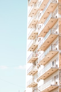 A high-rise building with multiple balconies featuring glass railings, bathed in bright sunlight against a clear sky.