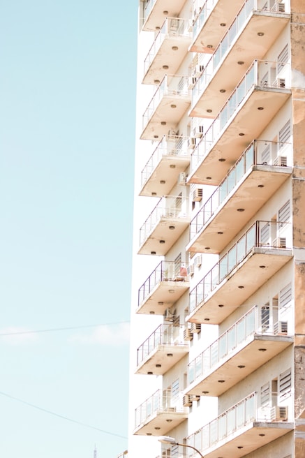 A high-rise building with multiple balconies featuring glass railings, bathed in bright sunlight against a clear sky.