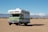A vintage-style RV is parked on a vast sandy desert landscape, with distant mountains under a clear blue sky.