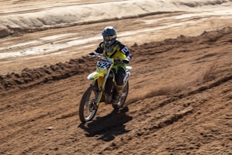 A person riding a motocross bike on a dirt track. The rider is wearing protective gear including a helmet, gloves, and a racing suit. The bike is kicking up dirt as it moves, and the surrounding area is a rough, barren landscape with tracks and grooves from other bikes.