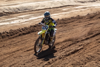 A person riding a motocross bike on a dirt track. The rider is wearing protective gear including a helmet, gloves, and a racing suit. The bike is kicking up dirt as it moves, and the surrounding area is a rough, barren landscape with tracks and grooves from other bikes.