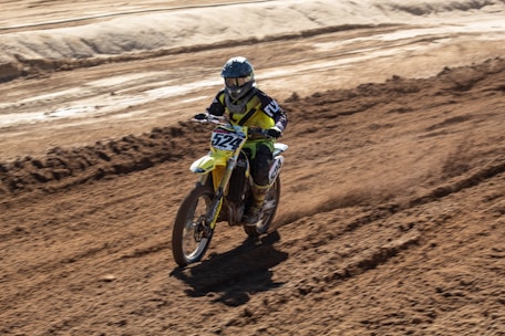 A person riding a motocross bike on a dirt track. The rider is wearing protective gear including a helmet, gloves, and a racing suit. The bike is kicking up dirt as it moves, and the surrounding area is a rough, barren landscape with tracks and grooves from other bikes.