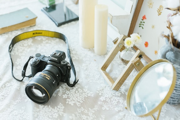 On a lace-covered surface, a Nikon camera with a yellow strap is featured prominently. Nearby are large ivory candles and a small vase containing white flowers on a wooden stand. A round mirror with a golden frame adds a reflective element. The background includes a hardcover book, a modern stand, and a board adorned with pressed flowers.
