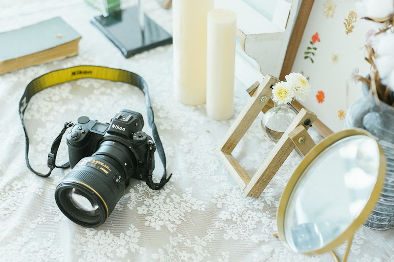 On a lace-covered surface, a Nikon camera with a yellow strap is featured prominently. Nearby are large ivory candles and a small vase containing white flowers on a wooden stand. A round mirror with a golden frame adds a reflective element. The background includes a hardcover book, a modern stand, and a board adorned with pressed flowers.