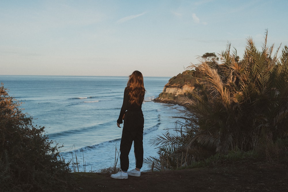 woman in black jacket standing on brown rock near body of water during daytime