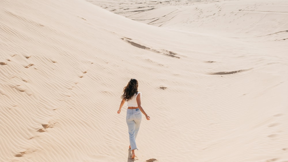 woman in white tank top and blue denim jeans walking on white sand during daytime