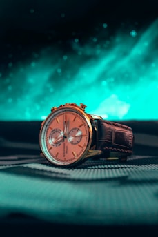 A close-up shot of a luxurious wristwatch with a gold casing and a brown leather strap. The watch face is rose gold with two subdials and elegant markers. The background features an abstract, blue-green, luminous design, adding a dynamic contrast to the watch.