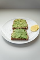 Two slices of bread topped with mashed avocado are placed on a white plate. The plate also contains two thin slices of lemon, adding a touch of freshness. There are small specks of black pepper sprinkled over the avocado toast. The background is a simple, light gray surface, giving a minimalist impression.