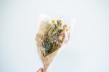 A bouquet of dried flowers wrapped in clear cellophane and brown paper. The arrangement includes various types and colors of flowers, such as yellow billy balls, white daisies, and pink statice. The bouquet is held up against a plain, light-colored background.